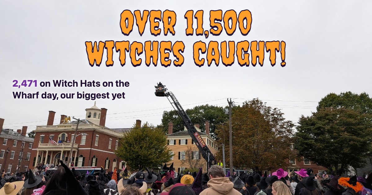 Over 11,500 Witches Caught! across the sky above a Salem fire department ladder truck, its bucket raising a photographer over a crowd in witch hats in front of the Custom House