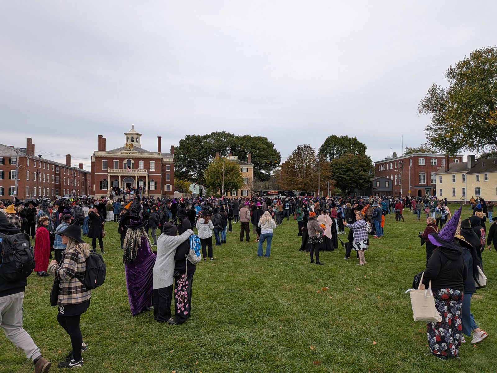 A lawn full of people in witch hats and capes milling about in front of the brick Custom House, with more arriving